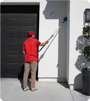 Person in a red shirt and cap painting an exterior white wall beside a garage door using a long-handled roller